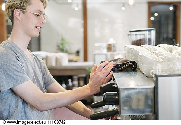 Young blond man with spectacles standing at an espresso machine in a coffee shop.