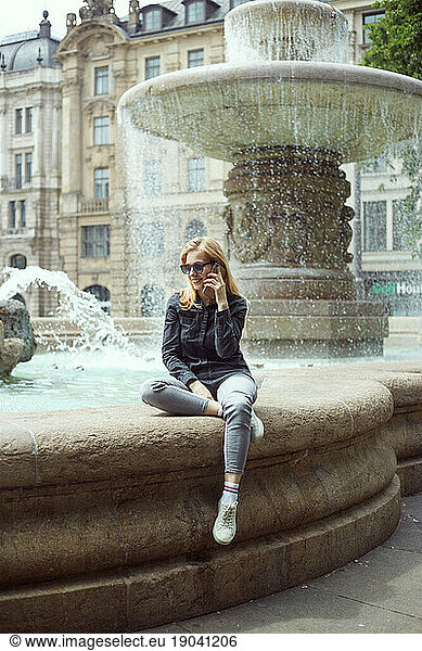 Young blond woman sitting by the fountain and talking on the phone