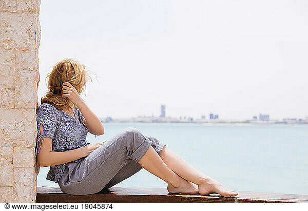 Young blond woman sitting on the fence and watching the sea