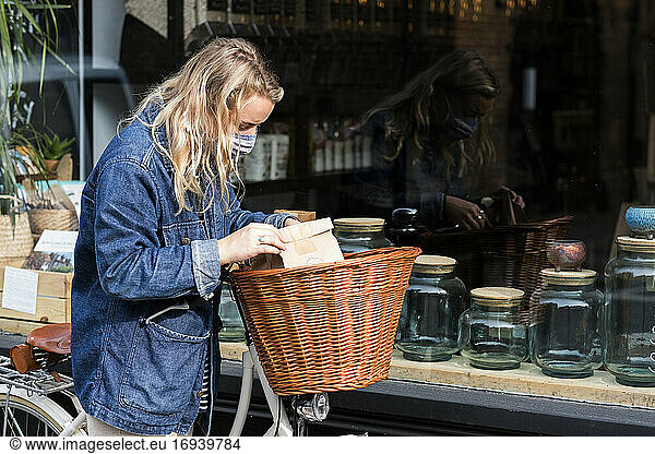 Young blond woman wearing face mask with bicycle  standing outside waste free wholefood store.