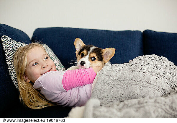 Young blonde girl snuggling small corgi puppy inside on blue couch
