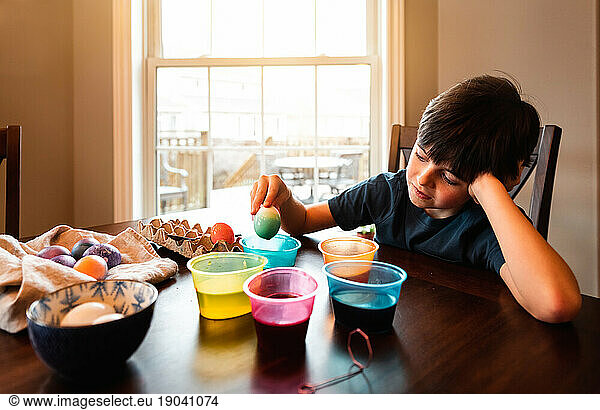 Young boy coloring Easter eggs with containers of dye at the table.