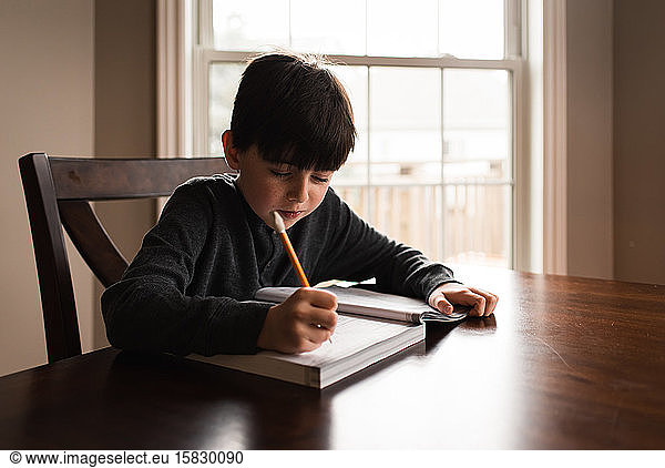 Young boy doing schoolwork in a workbook at home at the table.
