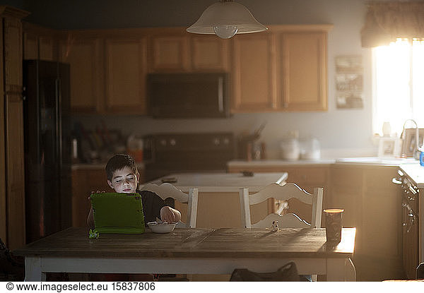 Young boy eats breakfast while watching his tablet in the kitchen