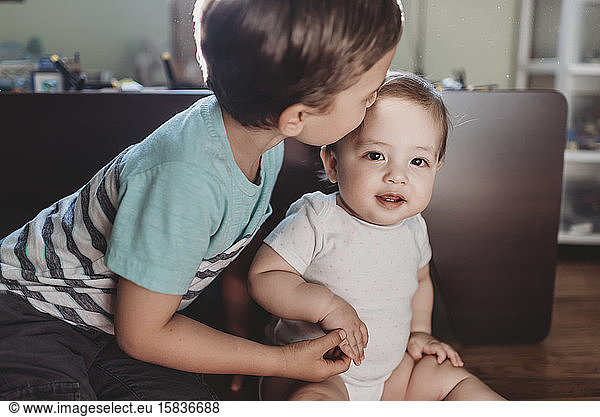 Young boy gently kissing baby sister and holding her tiny hand