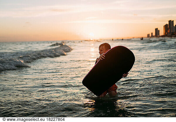Young boy holding board in ocean waves on beach at sunset