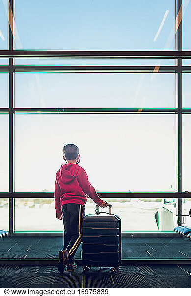 Young boy holding his suitcase looking out the window at airport.