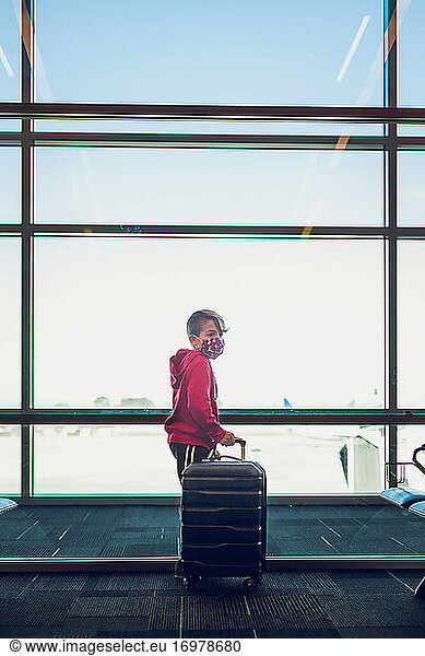 Young boy holding his suitcase standing next to window at airport.