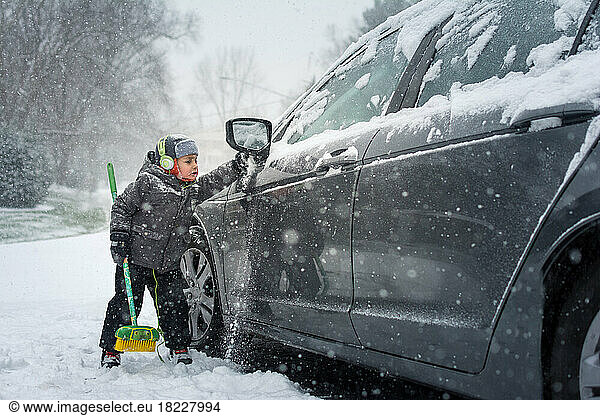 Young boy in gray jacket brushing snow off gray car on snowy day.