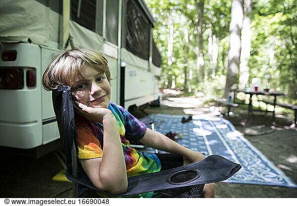 Young Boy in Tie Dye Shirt Sits in Camping Chair in Front of Pop Up