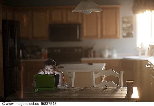 Young boy laughing while watching tablet at kitchen table eating food