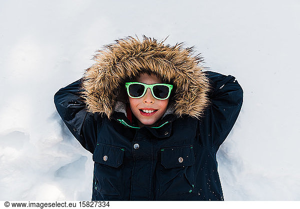 Young boy laying in snow with green sunglasses on Minnesota Spring day
