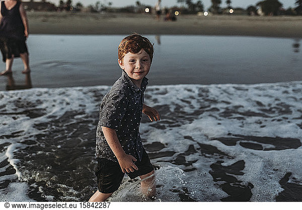 Young boy looking at camera while walking into the ocean at sunset