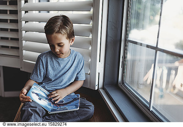 Young boy reading book by window with shutters
