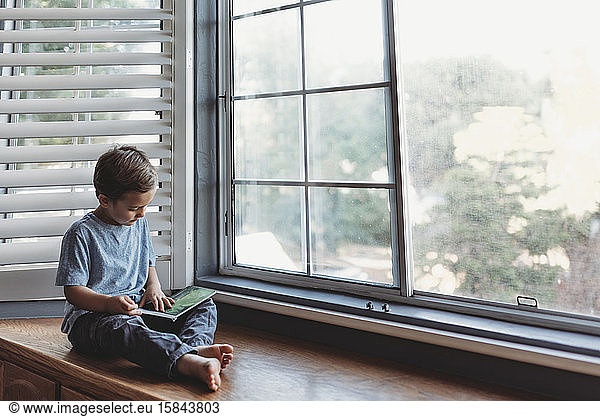 Young boy reads in window alcove with soft light and shutters