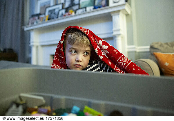 Young Boy Sits at Home Under a Red Winter Blanket Next to a Toy Bin