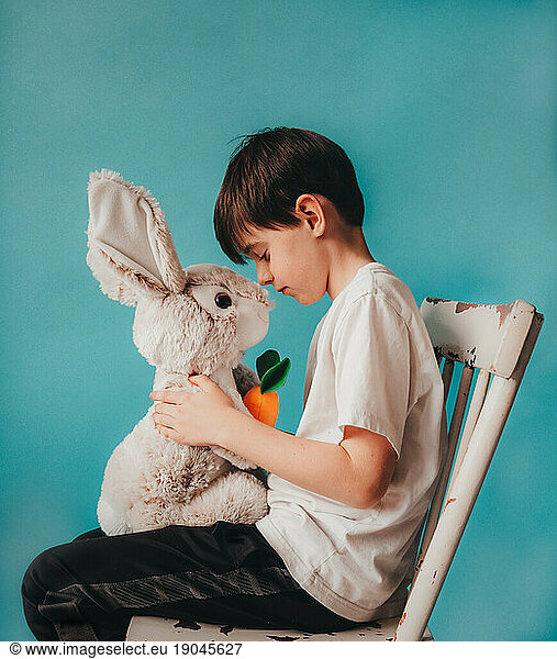Young boy sitting on chair touching noses with a stuffed bunny.