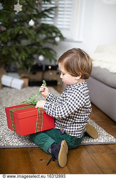 Young boy sitting on living room floor  unwrapping Christmas present in red box.