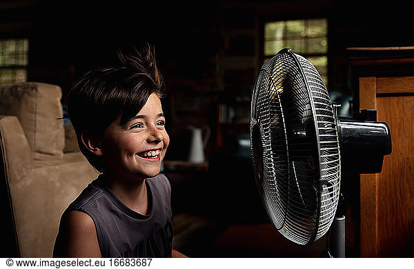 Young boy smiling as he is cooling off in front of a fan in dark room.