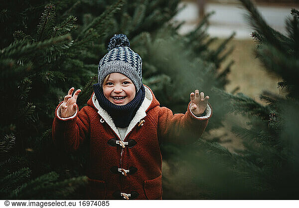 Young boy smiling between pine trees outside on cold winter day