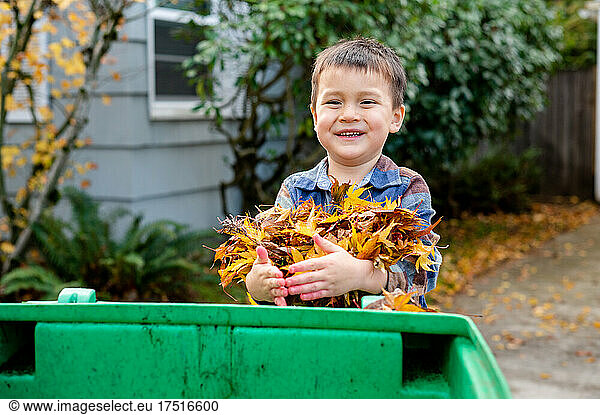 Young boy smiling with pile of leaves in arms and bin in front