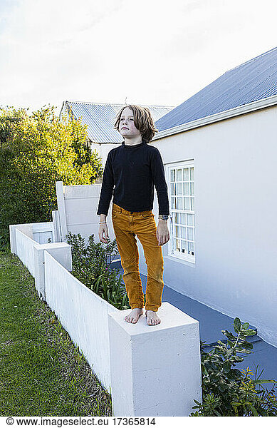 Young boy standing barefoot on a low wall outside a house