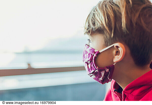 Young boy wearing a mask looking out the window at airport.