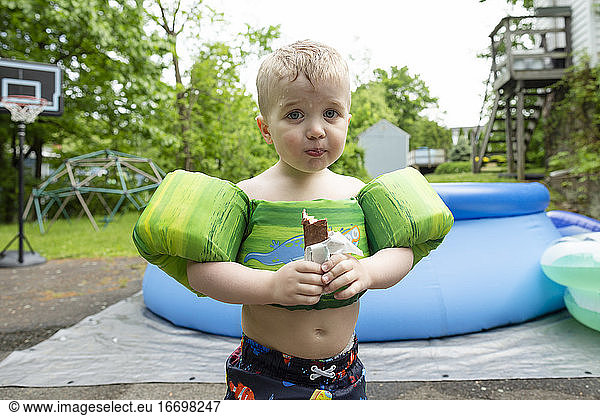 Young boy wearing swimming gear stands eating candy in yard with pool