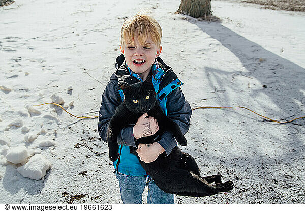 Young boy wearing winter coat carries heavy black cat looking in