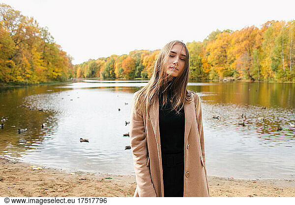 Young brunette girl enjoying autumn lakeside in camel coat in park