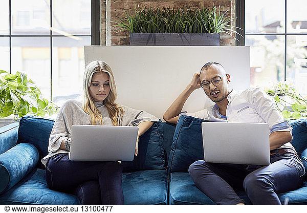 Young business people using laptops while sitting on sofa at creative office