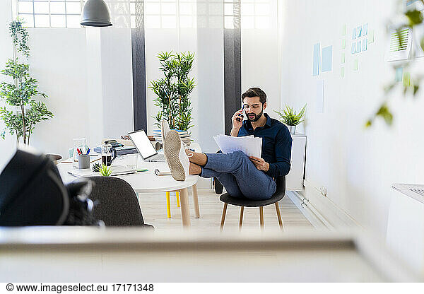 Young businessman discussing on smart phone while sitting with feet up at desk in office