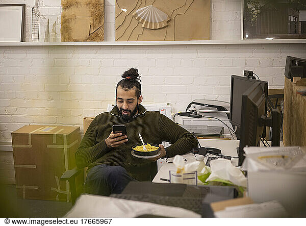 Young businessman eating lunch and using smart phone in office
