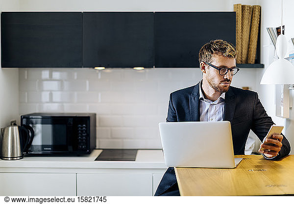 Young businessman using laptop and cell phone in kitchen