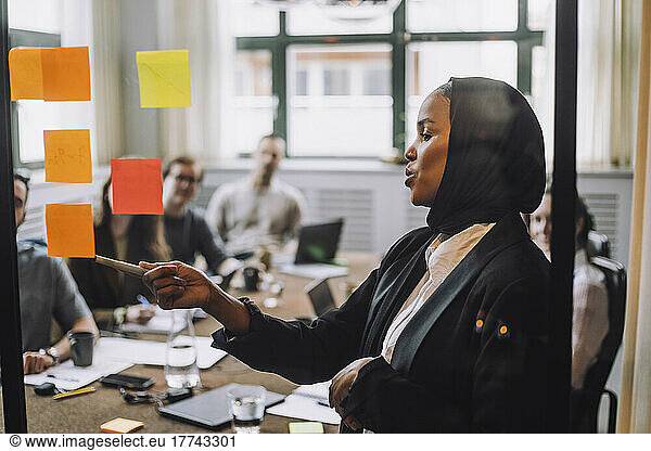 Young businessman wearing headscarf explaining adhesive notes to colleagues during meeting in creative office