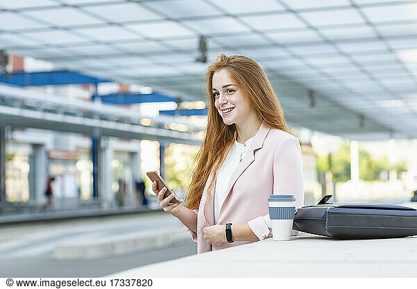 Young businesswoman looking away while holding smart phone at railway station