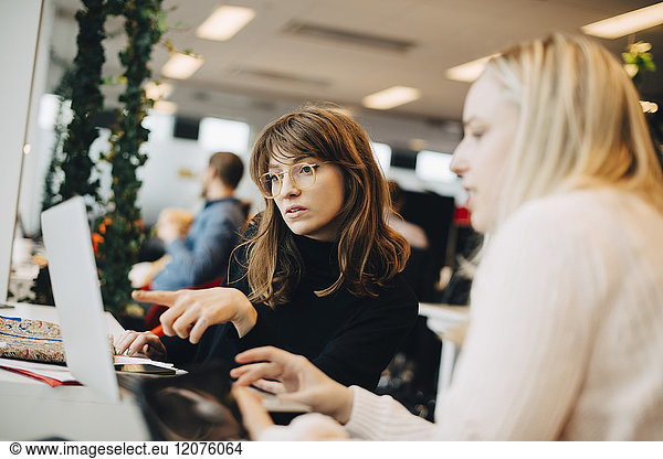 Young businesswoman pointing at laptop to female colleague sitting in office