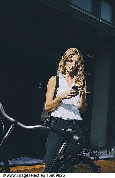 Young businesswoman using mobile phone while standing with bicycle in city