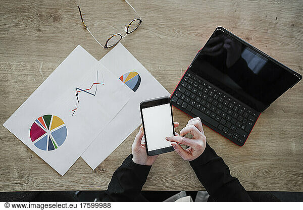 Young businesswoman using smart phone by laptop and documents on desk in office