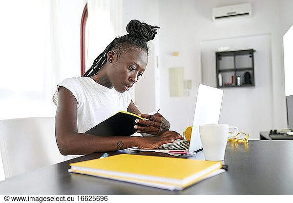 Young businesswoman with book using laptop on desk while sitting in home office