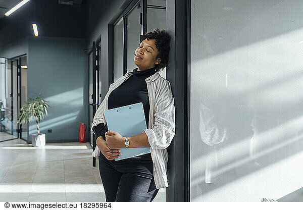 Young businesswoman with eyes closed leaning on glass in office