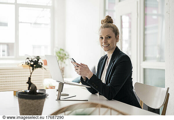Young businesswoman with smart phone over laptop sitting at desk in office