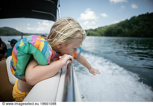 Young child leaning over boat railing to touch wake on lake