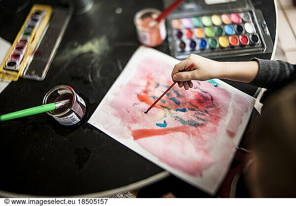 young child's hand painting with a brush surrounded by art supplies