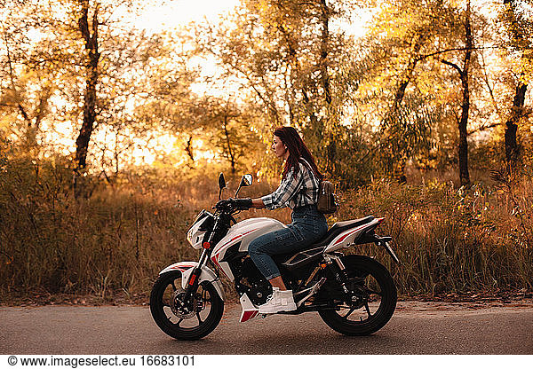 Young confident woman riding motorcycle on country road at sunset
