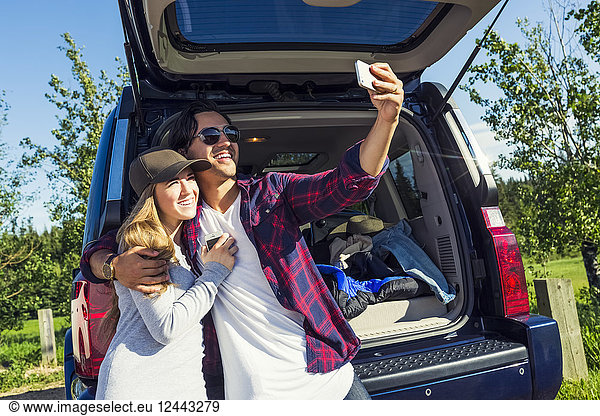 Young couple at their vehicle with the back open taking a self-portrait with their cell phone  Edmonton  Alberta  Canada