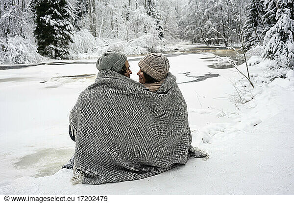 Young couple covered in blanket looking at each other while sitting by frozen river in forest