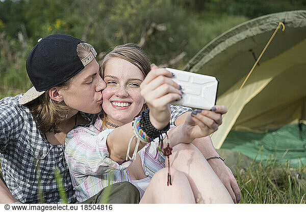 Young couple kissing and taking selfie with smart phone in front of camp tent  Bavaria  Germany