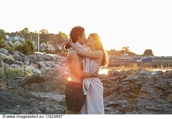 Young couple kissing while standing on rock at seashore during sunset