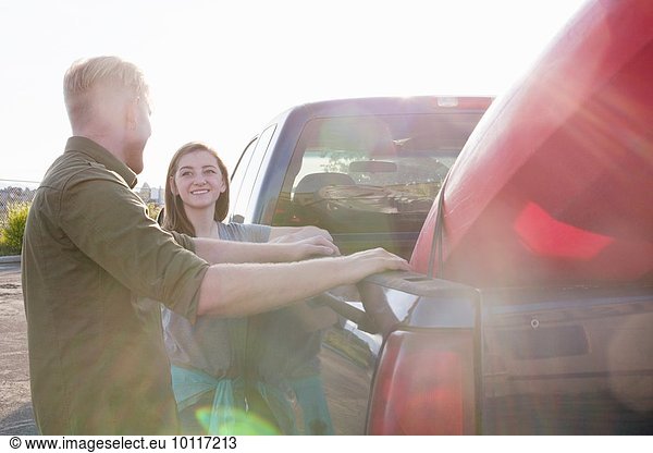 Young couple leaning against pick up truck  looking at each other smiling  lens flare